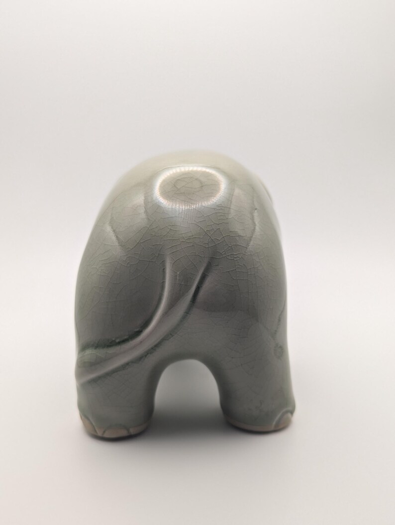 May include: A light grey-green ceramic elephant figurine, seen from behind. The elephant has a textured, crackled surface and a curled tail. The figurine has a rounded body and stands on four legs. The measurements are in centimetres.