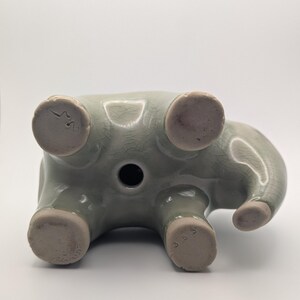 May include: A light green ceramic elephant figurine, seen from below. The elephant has a trunk, four legs, and a circular hole. The feet are a lighter colour, and the figurine has a crackled texture. Measurements are in centimetres.