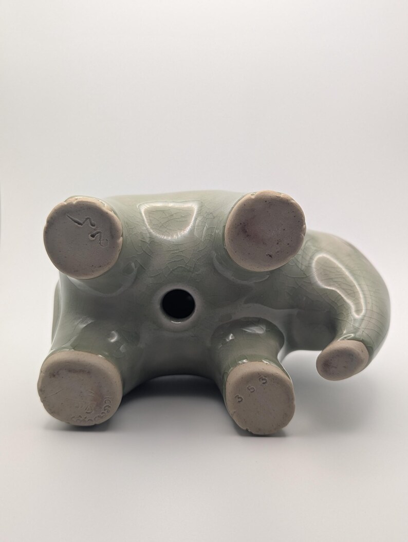 May include: A light green ceramic elephant figurine, seen from below. The elephant has a trunk, four legs, and a circular hole. The feet are a lighter colour, and the figurine has a crackled texture. Measurements are in centimetres.