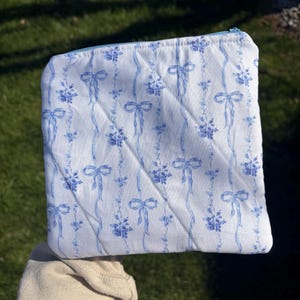 Può includere: Un astuccio trapuntato bianco con un motivo floreale e a nastro blu. L'astuccio ha una cerniera blu chiaro e un design trapuntato diagonale. Il motivo presenta fiocchi blu e accenti floreali.