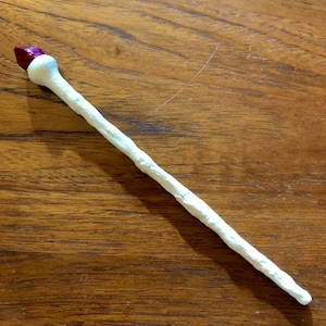 May include: A white wand with a textured surface and a red, gem-like tip. The wand is lying on a wooden surface. The wand is approximately 12 inches long.