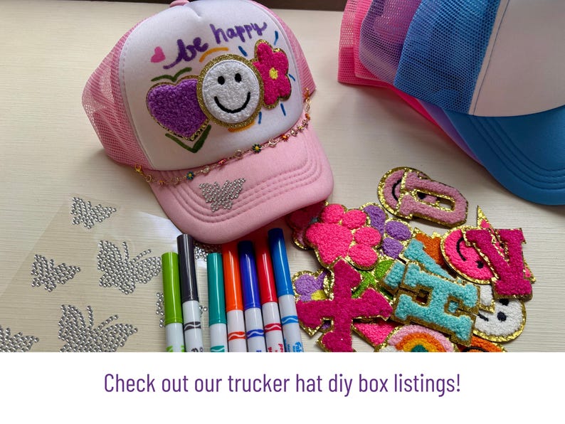 May include: Pink trucker hat with a white mesh back and a curved brim. The hat features a smiley face, heart, and flower design. Also shown are colorful markers, patches, and rhinestone butterfly stickers. The text "be happy" is on the front.