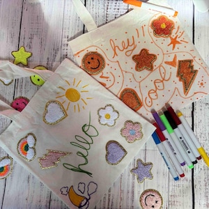 Puede incluir: Dos bolsas de lona blancas con parches y dibujos coloridos. Una bolsa dice "hey! cool" y la otra dice "hello". Ambas bolsas est&aacute;n decoradas con parches y dibujos coloridos. Tambi&eacute;n se incluye un juego de marcadores de colores.