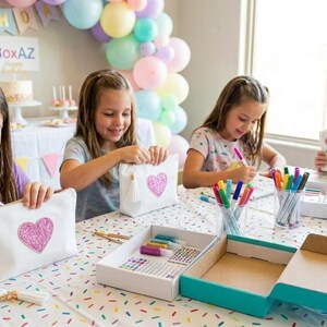 May include: Four young girls are gathered around a table, engaged in a craft activity. They are decorating white canvas bags with pink heart designs using markers. The table is covered with craft supplies, including markers, stickers, and paint.