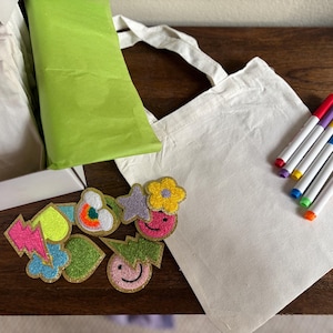 Puede incluir: Una bolsa de tela blanca con un juego de marcadores de colores y una variedad de parches termoadhesivos de colores. Los parches tienen forma de rayo, coraz&oacute;n, nube, estrella, flor y cara sonriente.