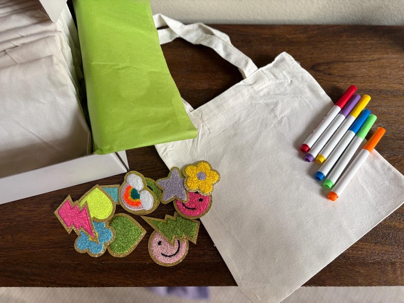 Puede incluir: Una bolsa de tela blanca con un juego de marcadores de colores y una variedad de parches termoadhesivos de colores. Los parches tienen forma de rayo, coraz&oacute;n, nube, estrella, flor y cara sonriente.