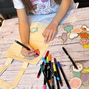 Puede incluir: Una persona est&aacute; personalizando una bolsa de lona blanca con marcadores de colores. La bolsa tiene varios parches coloridos pegados, incluyendo una flor, un coraz&oacute;n, un rayo y una cara sonriente.