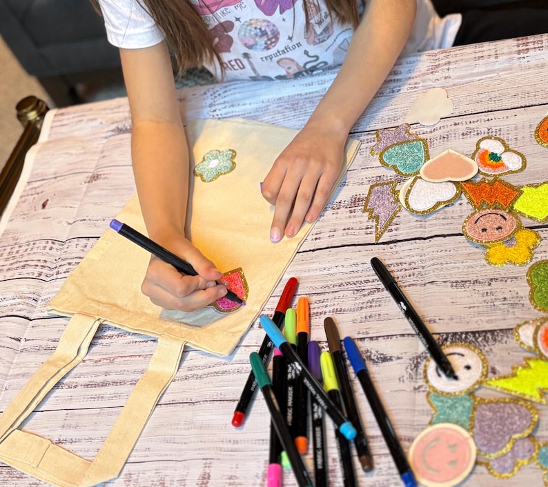 Puede incluir: Una persona est&aacute; personalizando una bolsa de lona blanca con marcadores de colores. La bolsa tiene varios parches coloridos pegados, incluyendo una flor, un coraz&oacute;n, un rayo y una cara sonriente.