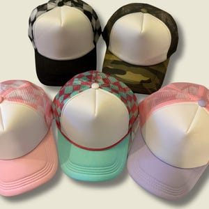 May include: Five trucker hats with white foam fronts and mesh backs. The hats feature various patterns and colors, including black and white checkered, camouflage, pink, teal, and lavender. Each hat has a curved brim and a button on top.