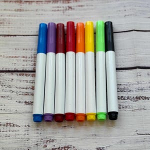 May include: A collection of eight colorful markers arranged on a light wood-grain surface. The markers have white barrels and caps in blue, purple, maroon, red, orange, yellow, green, and black. The markers are ideal for art projects.