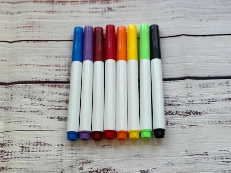 May include: A collection of eight colorful markers arranged on a light wood-grain surface. The markers have white barrels and caps in blue, purple, maroon, red, orange, yellow, green, and black. The markers are ideal for art projects.