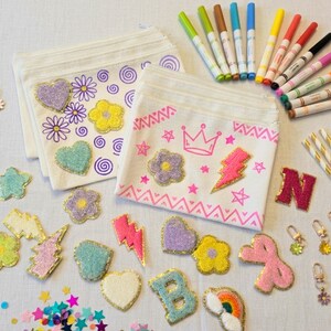 May include: An assortment of craft supplies including two white fabric pouches decorated with embroidered flowers, hearts, and swirls. Also shown are colorful markers, glittery patches, keychains, and gold-striped straws. A creative and colorful craft kit.