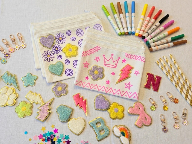 May include: An assortment of craft supplies including two white fabric pouches decorated with embroidered flowers, hearts, and swirls. Also shown are colorful markers, glittery patches, keychains, and gold-striped straws. A creative and colorful craft kit.