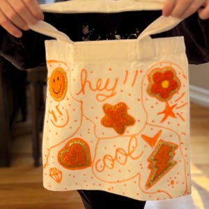 May include: Off-white tote bag with orange illustrations and text. The bag features a smiling face, star, heart, flower, and lightning bolt designs. The words "hey!!" and "cool" are written in orange script.
