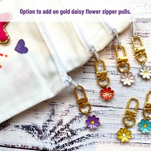 May include: White canvas zipper pouches with gold daisy flower zipper pulls in various colors. The image shows the option to add the daisy flower zipper pulls. The daisy flowers are in red, pink, white, purple, yellow, and blue.