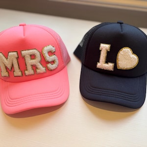 May include: Two trucker hats. One is pink with "MRS" in white and gold letters. The other is navy blue with a white "L" and a white heart with a gold border. Both hats have a mesh back.