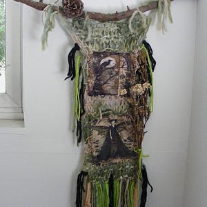 May include: A wall hanging featuring a thick, sage green knitted fiber base suspended from a natural wooden branch. It includes two square fabric patches with dark illustrations, black and green yarn fringe, a pinecone, and dried floral stems.