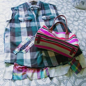 May include: A sleeveless button-down shirt featuring a green, white, and dark gray plaid pattern with a frayed, multi-colored plaid hem. A horizontal striped handbag in shades of pink, white, and black with brown leather handles rests on top.
