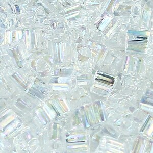 May include: A collection of small, clear, cylindrical glass beads with an iridescent finish. Each bead features a central hole and a reflective surface that displays subtle rainbow-like highlights when light hits the faceted, transparent glass material.