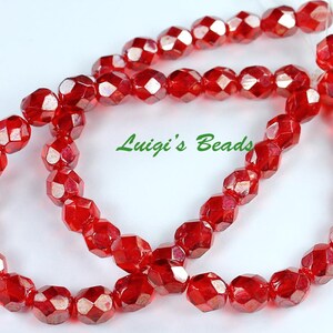 May include: A strand of faceted, round, translucent red glass beads arranged in a circular loop. The beads have a reflective, shiny surface. The text Luigi's Beads is printed in green in the center of the loop of red beads.