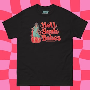 May include: A black short-sleeved t-shirt featuring a graphic of a figure in a light blue dress standing in red flames. The text reads Hell Yeah Babes in a red font. The shirt has a small logo on the inside neck area that reads Celestial Skulls.