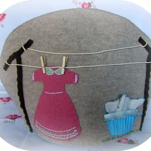 May include: A gray felt decorative pillow with a whimsical laundry scene. A maroon dress hangs on a clothesline between two black posts. A blue basket holds folded white fabric. The pillow is on a floral patterned surface.