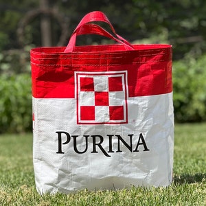 May include: A red and white Purina tote bag with red handles. The bag features the Purina logo and a checkered pattern. The bag is made of a durable material and is designed for carrying items.