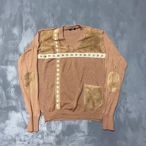 Peut inclure: Un pull col en V beige avec des empiècements en suédine marron sur les coudes et une poche assortie sur le côté inférieur droit. Le devant est orné d'une bande à motif quadrillé crème disposée verticalement et horizontalement.