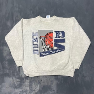 Pode incluir: Camisola cinza claro com um gráfico de basquete. O design inclui uma bola de basquete vermelha, uma rede e as palavras "DUKE" em azul. As palavras "Blue Devils" estão em uma faixa azul. Roupa casual.