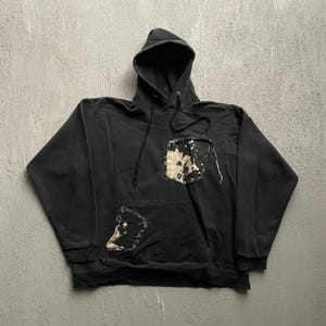 Peut inclure: Un sweat-shirt à capuche noir avec des manches longues, un cordon de serrage et deux empiècements rectangulaires présentant un motif délavé, tacheté de blanc et de noir. Un empiècement est situé sur la poitrine et l'autre sur la poche avant.