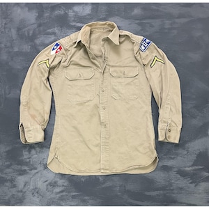 May include: Tan long-sleeved shirt with two chest pockets and button closures. Patches with insignia are on the shoulders. The shirt has a classic military style.