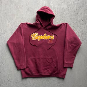 Puede incluir: Una sudadera con capucha de color granate que presenta la palabra Gophers en letra cursiva amarilla con un borde blanco, la palabra Minnesota en texto amarillo pequeño y un logotipo M en granate y amarillo. Incluye bolsillo frontal y cordones.