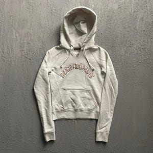 May include: An off-white long-sleeve hooded sweatshirt featuring a front kangaroo pocket and a hood with adjustable drawstrings. The word ABERCROMBIE is embroidered in a curved, arched design across the chest in a light brown thread colour.