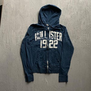 May include: A dark blue zip-up hoodie featuring a white distressed graphic on the chest that reads HOLLISTER 1922. The garment includes a hood with matching drawstrings, long sleeves with ribbed cuffs, and two front pockets, all in a soft fabric.