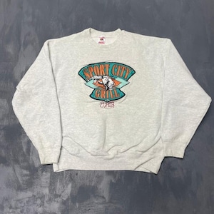 Op de afbeelding: Een lichtgrijs sweatshirt met een ronde hals. De voorkant heeft een kleurrijke afbeelding met de tekst "SPORT CITY GRILL" in teal en oranje. De afbeelding bevat een illustratie van een chef-kok en een grill.