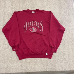 May include: A dark red long-sleeve crewneck sweatshirt featuring the text 49ERS in a stylised white and red font across the chest, with the San Francisco 49ers team logo centred below the text and a small rectangular brand tag on the lower left hem.