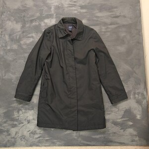 May include: A dark grey GAP raincoat with a collared neckline and long sleeves. The coat is buttoned up and appears to be made of a lightweight material. The GAP logo is visible on the collar.