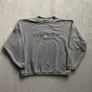 Könnte beinhalten: Ein graues langärmeliges Sweatshirt mit Rundhalsausschnitt und einem zentralen Aufdruck mit dem Text Genuine Mickey Trademark sowie einer kleinen Mickey-Mouse-Illustration. Das Kleidungsstück hat gerippte Bündchen und einen gerippten Saum.
