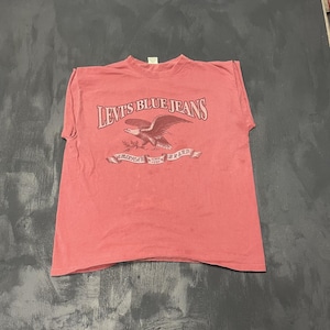 May include: A faded red t-shirt with the words "Levi's Blue Jeans" and an eagle graphic. Underneath is a banner reading "America's Brand Since 1850". The shirt has a crew neck and short sleeves.