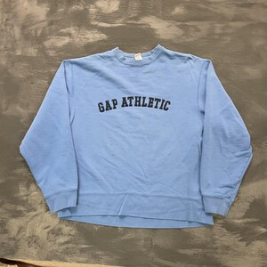 以下が含まれることがあります： ライトブルーのGAP Athleticスウェットシャツ。クルーネックのスウェットシャツは長袖で、裾と袖口はリブ編みです。「GAP ATHLETIC」の文字が胸に黒でプリントされています。スウェットシャツは柔らかく快適な素材で作られています。