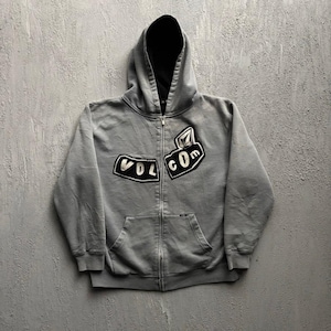 May include: A grey zip-up hoodie featuring a black interior hood lining and two black patches on the chest with white embroidered text reading VOLCOM. The garment includes a full-length front zip, long sleeves, and a front kangaroo pocket.