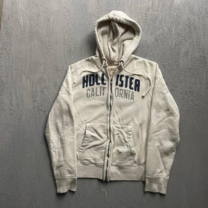 May include: A cream-colored zip-up hoodie featuring a hood with drawstrings and a waffle-knit lining. The front displays the dark blue embroidered text HOLLISTER and the grey embroidered text CALIFORNIA, along with two front pouch pockets.