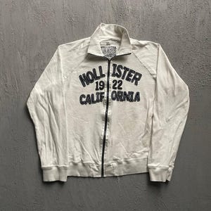 May include: A white long-sleeve zip-up jacket with a high collar and black text on the chest that reads HOLLISTER 19 22 CALIFORNIA. The garment features a central black zipper, ribbed cuffs, and a size XL label printed on the inner neck area.