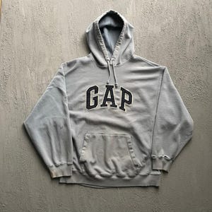 May include: A light gray hooded sweatshirt featuring the word GAP in dark navy blue block letters across the chest. The garment includes a front kangaroo pocket, a drawstring hood, and ribbed cuffs with visible signs of wear and fraying at the edges.