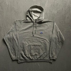 Può includere: Una felpa con cappuccio a maniche lunghe in grigio mélange, dotata di una tasca a marsupio frontale e di un piccolo logo ricamato in blu e bianco sul petto sinistro. La felpa presenta un cordino al collo e polsini e orlo a coste.