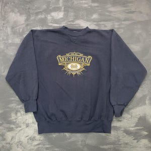 May include: Navy blue sweatshirt with a crew neck. The front features a gold and yellow emblem with the word "MICHIGAN" and the text "WOLVERINES". The sweatshirt is made of a soft, comfortable material.