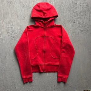 Op de afbeelding: Een felrode hoodie met ritssluiting, een volledige rits aan de voorzijde, een capuchon met een klein geborduurd logo op de voorzijde en gebogen naaddetails over de borst en romp. Het kledingstuk heeft lange mouwen en een geribbelde zoom.