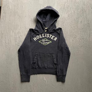 May include: A dark navy blue hooded sweatshirt featuring a front kangaroo pocket and long sleeves. The chest displays the white embroidered text HOLLISTER above a green diamond-shaped outline containing the white script text Wave Champs.