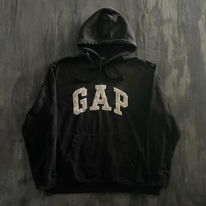May include: A black long-sleeve hooded sweatshirt featuring the word GAP in large, light-coloured block letters across the chest. The garment includes a drawstring hood and a large front kangaroo pocket, constructed from a solid black fabric.