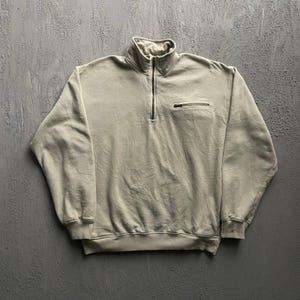 May include: A light beige long-sleeve pullover sweatshirt featuring a quarter-length zipper at the collar and a small horizontal zippered pocket on the upper left chest. The garment has ribbed cuffs and a ribbed hem, with a stand-up collar design.